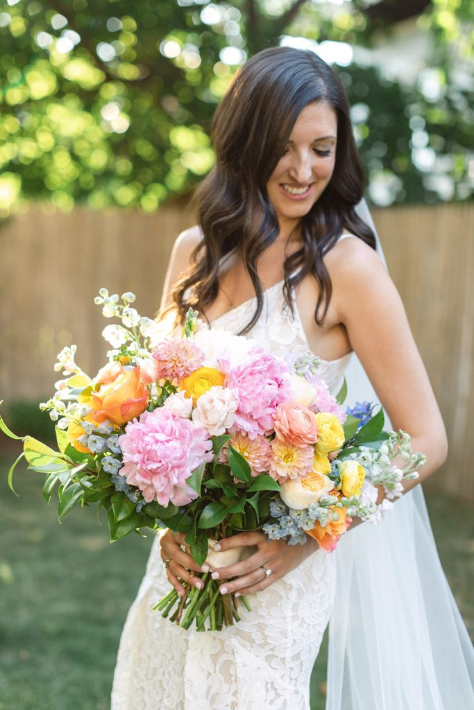 A bride looks down, smiling, at her orange, pink and white bouquet