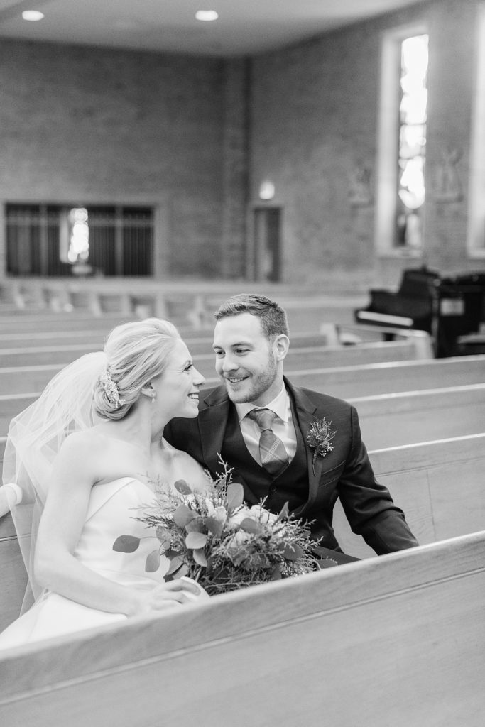 A bride and groom smile, sitting next to each other in a church pew